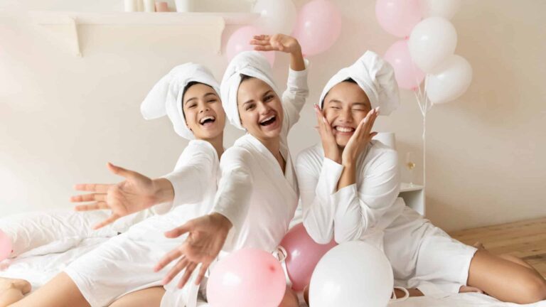Three people wearing white bathrobes and towels on their heads sit on a bed surrounded by pink and white balloons, smiling and posing for the camera.