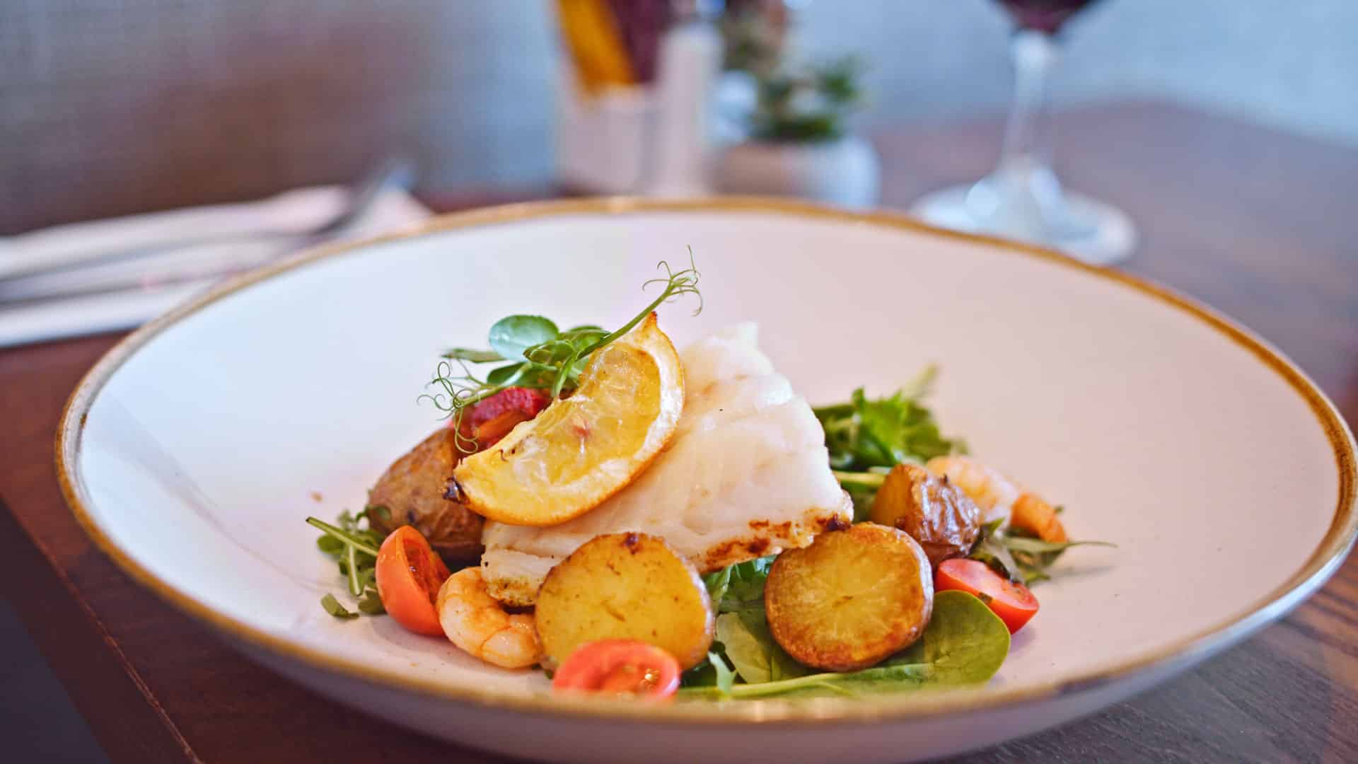 Pan-fried hake topped with lemon slice, served with cherry tomatoes, prawns, baby potatoes, and mixed greens on a wide white plate.