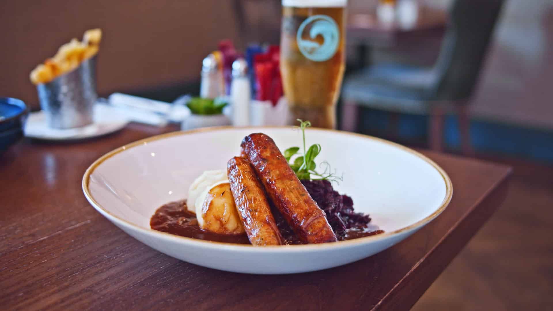 Traditional bangers and mash with gravy, red cabbage, and greens served in a white bowl with a glass of beer in the background.