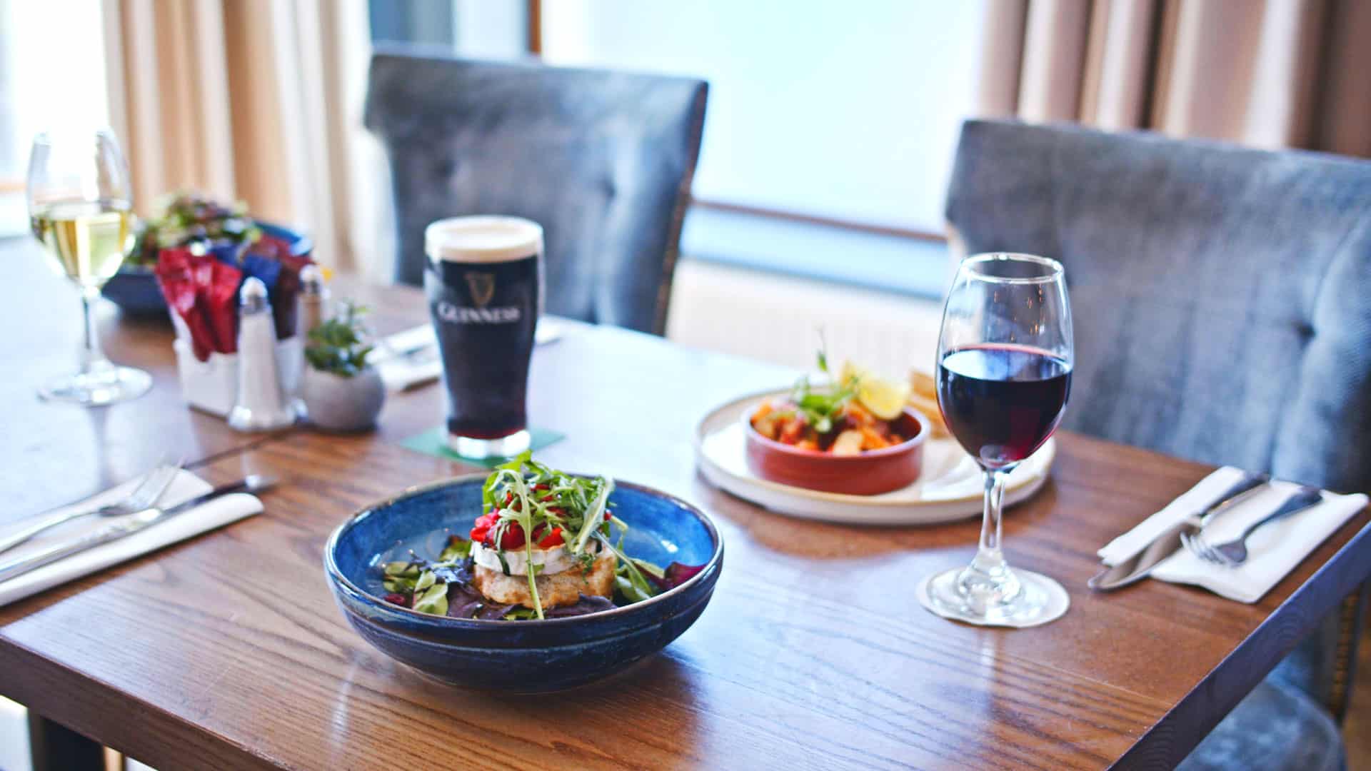 A duo of plated dishes including fish and chips and a pasta bowl, with glasses of red wine, white wine, Guinness, and water on a cosy table for two