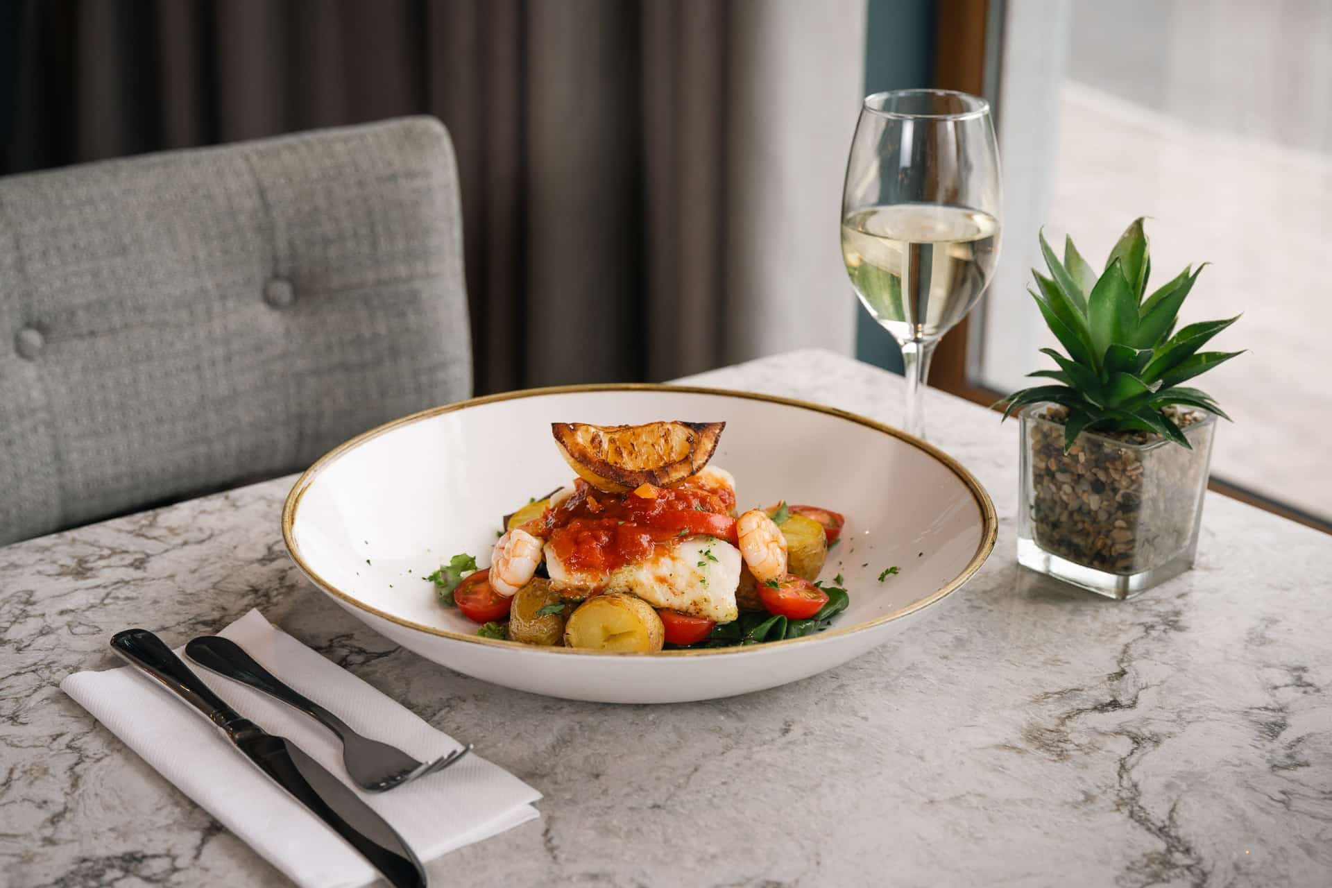 Garlic and chilli prawns served over roast baby potatoes, wilted spinach, and cherry tomatoes, topped with tomato sauce and grilled lemon, with a glass of white wine on a marble table.